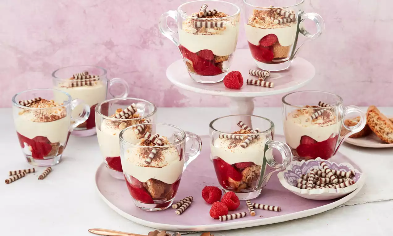 Dessert aus heller Creme auf geschichtetem roten Fruchtpüree, garniert mit Schokospänen in Glastassen auf rosa Platte mit Himbeeren