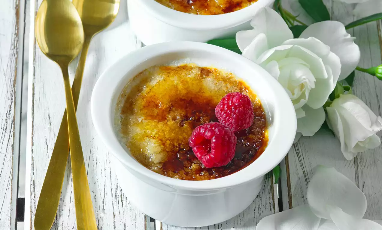 Crème brûlée s karamelově zlatavou krustou nalomenou uprostřed, ozdobené sytě červenými malinami v bílém keramickém ramekinu na dřevěném stole