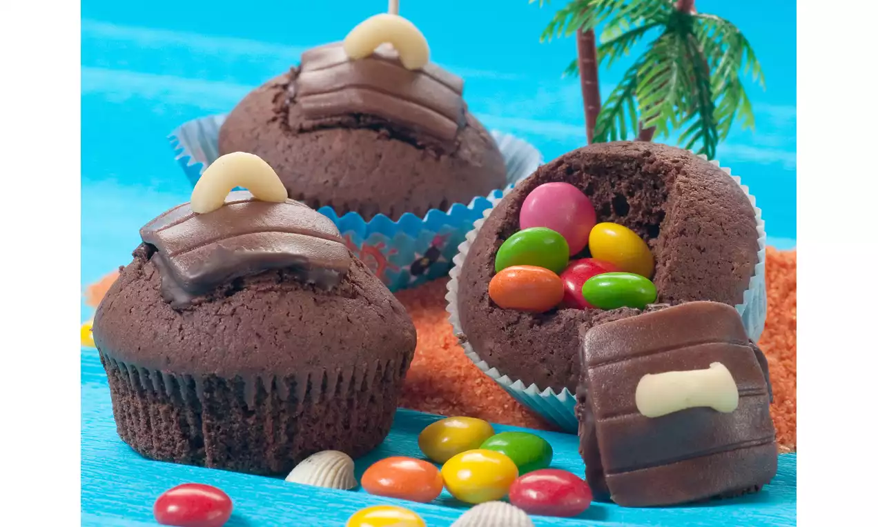 Schokoladen-Muffins in blauen Förmchen mit aufgesetzten Schokodeckeln, gefüllt mit bunten Schokolinsen, arrangiert auf blauem Untergrund mit Sand und Palmen-Deko