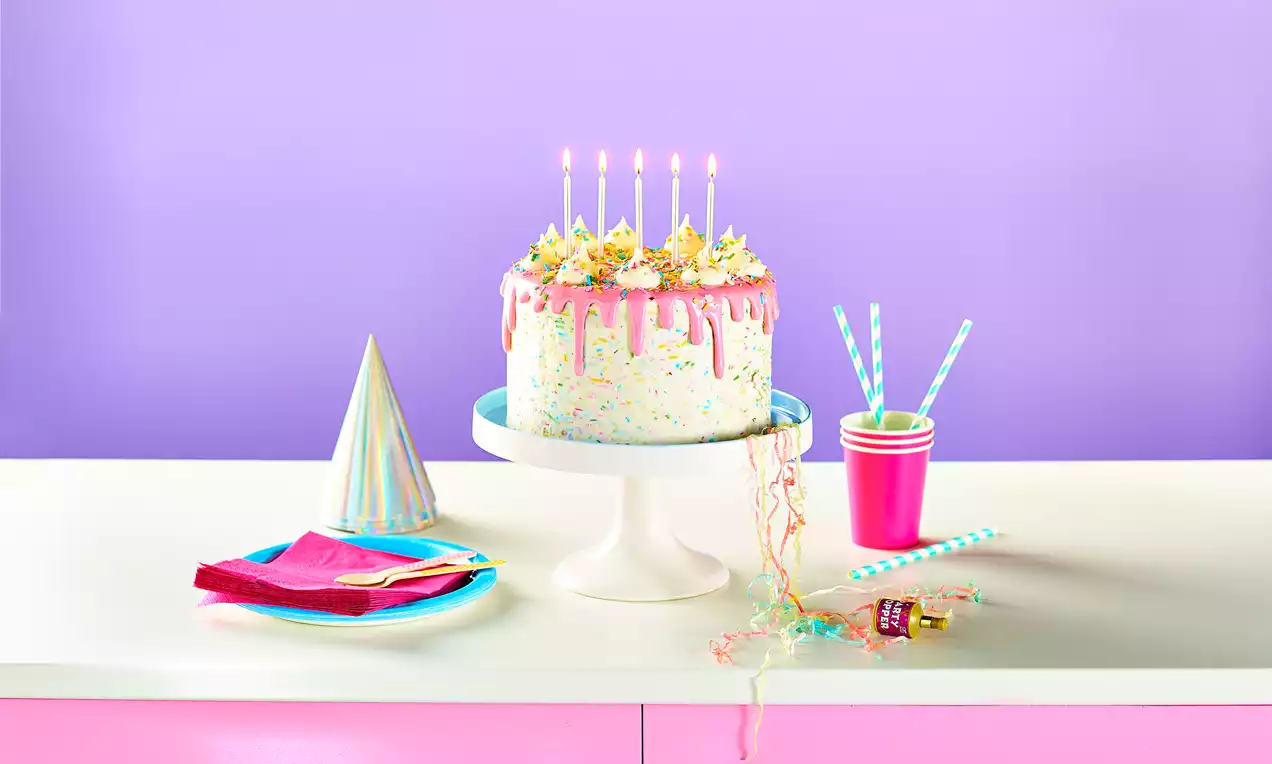 A round, sprinkle-speckled cake with pink drips, yellow frosting swirls, and lit candles on a white stand against a pastel backdrop