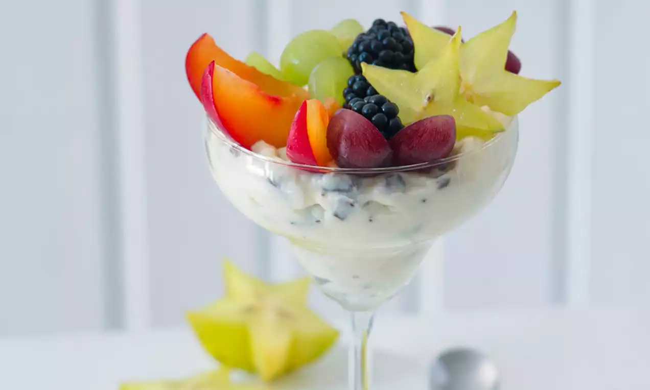 Sklenice naplněná světlým jogurtem s kousky, ozdobená švestkami, hrozny, ostružinami a plátky karamboly na světlém podkladu