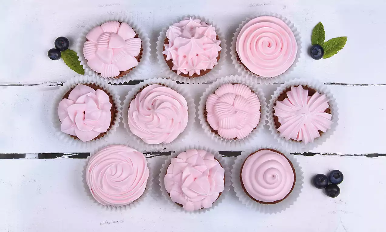 Schoko-Muffins mit zartrosa Cremehauben in verschiedenen Mustern, serviert in Papierförmchen, dekoriert mit Blaubeeren und Minzblättern auf weißem Holztisch