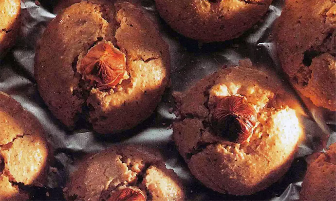 Ronde, goudbruine notenkoekjes met een gebarsten textuur en donkere hazelnoten in het midden, gelegd op een metallic ondergrond