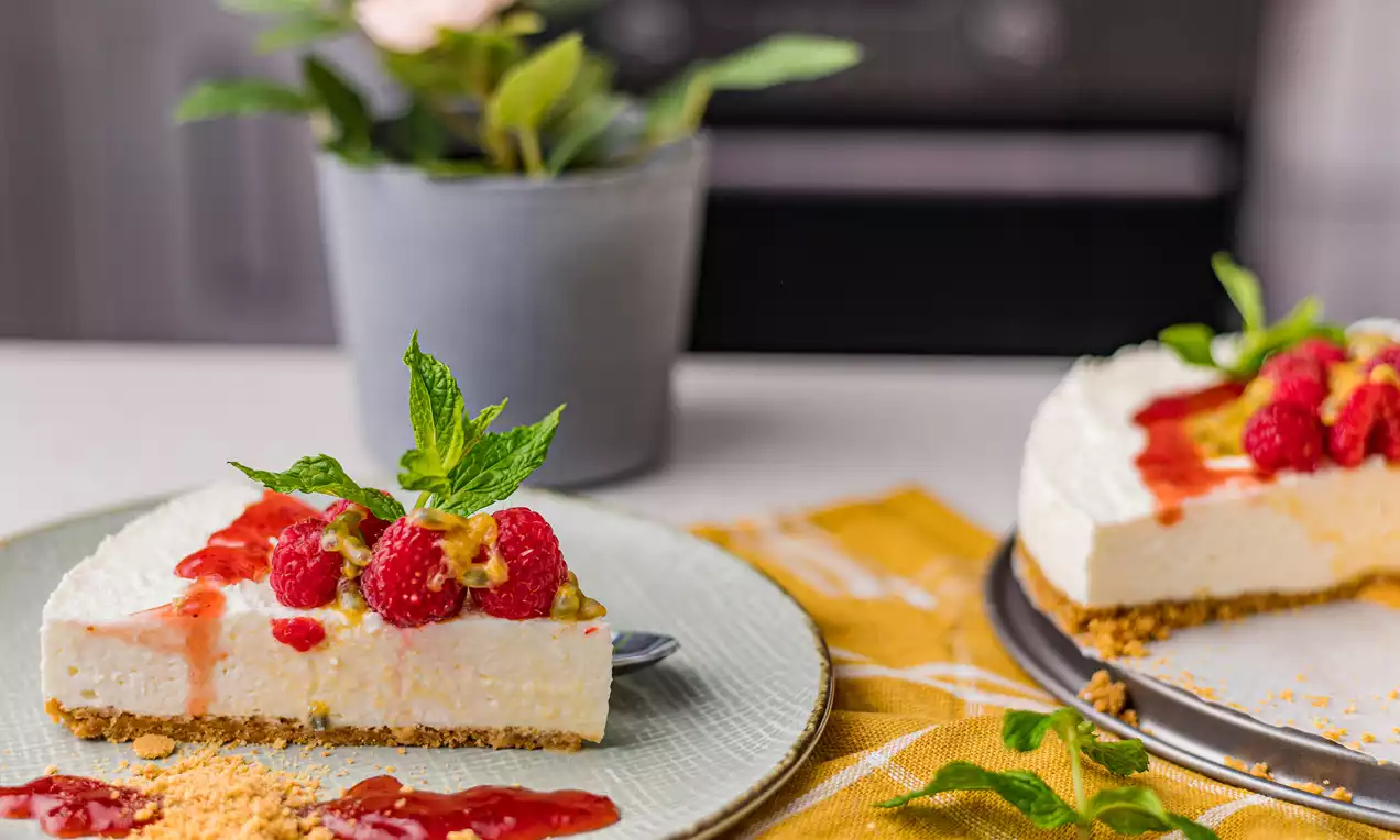 Cheesecake de vanilie fără coacere cu textură fină și strat crocant de biscuiți, decorat cu zmeură, sos roșu și frunze proaspete de mentă pe o farfurie albă