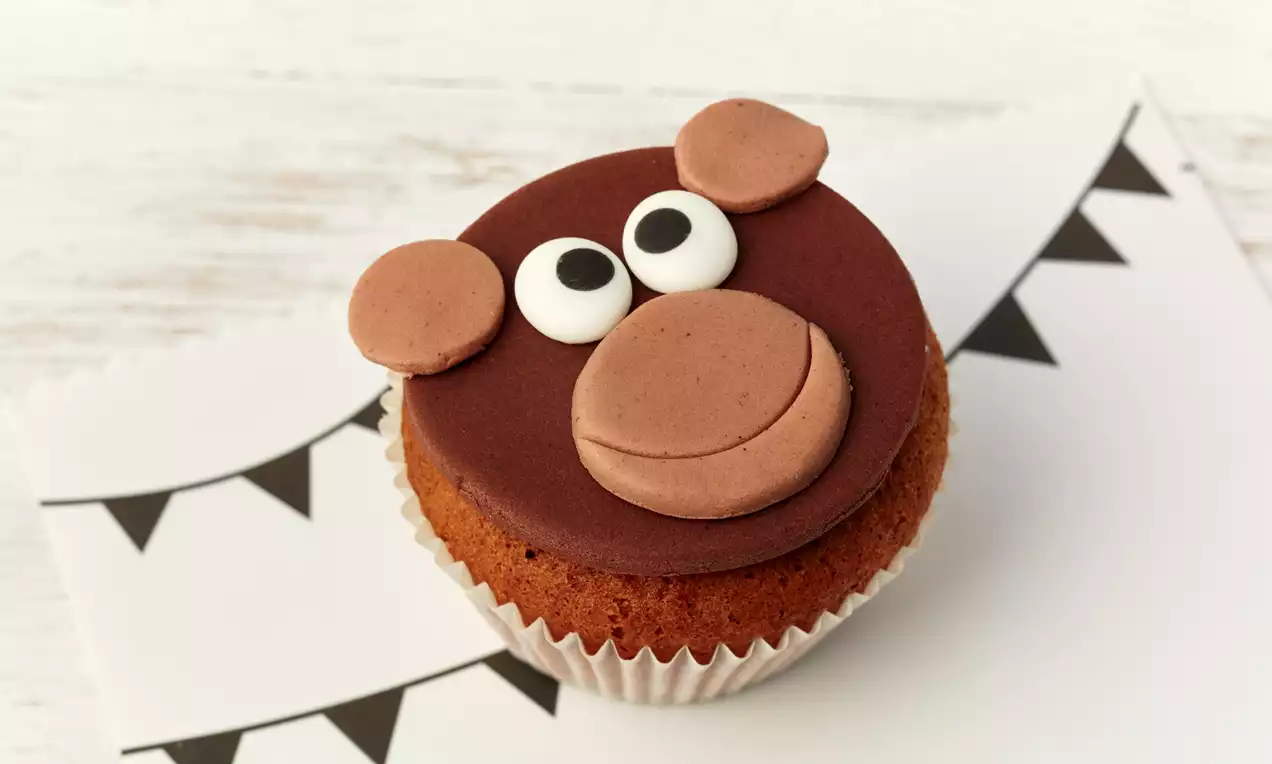 Brauner Cupcake mit einem flachen Schokomantel, Zuckeraugen, hellbrauner Schnauze und Ohren, arrangiert auf weißem Papier mit schwarzen Dreiecken