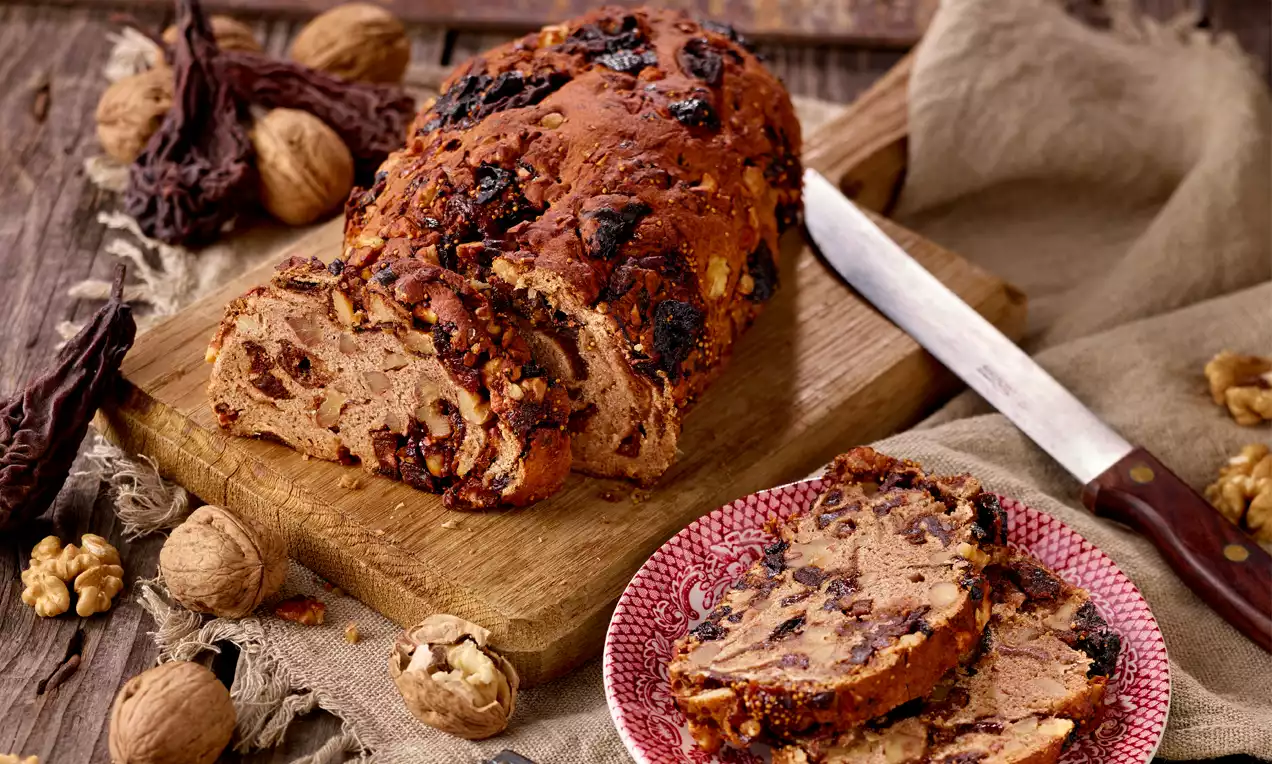 Kletzenbrot mit goldbrauner Kruste, gefüllt mit dunklen Trockenfrüchten und Nüssen, angeschnitten auf Holzbrett neben roten Teller und Messer