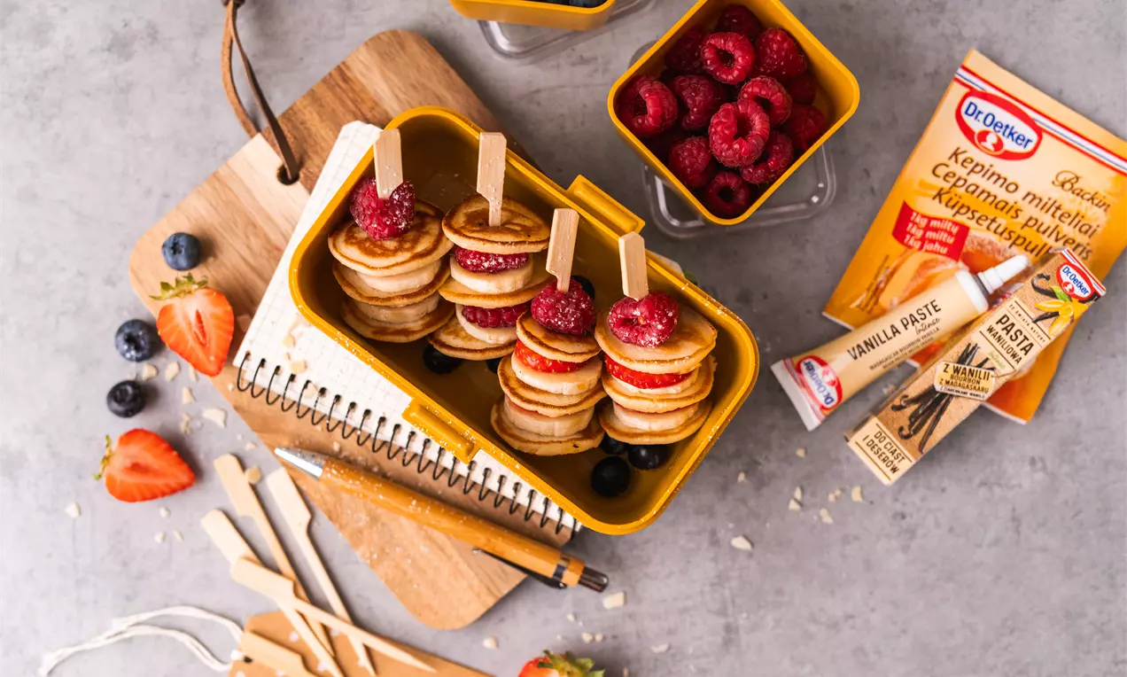 Auksiniai mini blyneliai suapvalintos formos, suverti ant iešmelių, dekoruoti avietėmis ir pateikti geltoname dubenėlyje ant pjaustymo lentelės