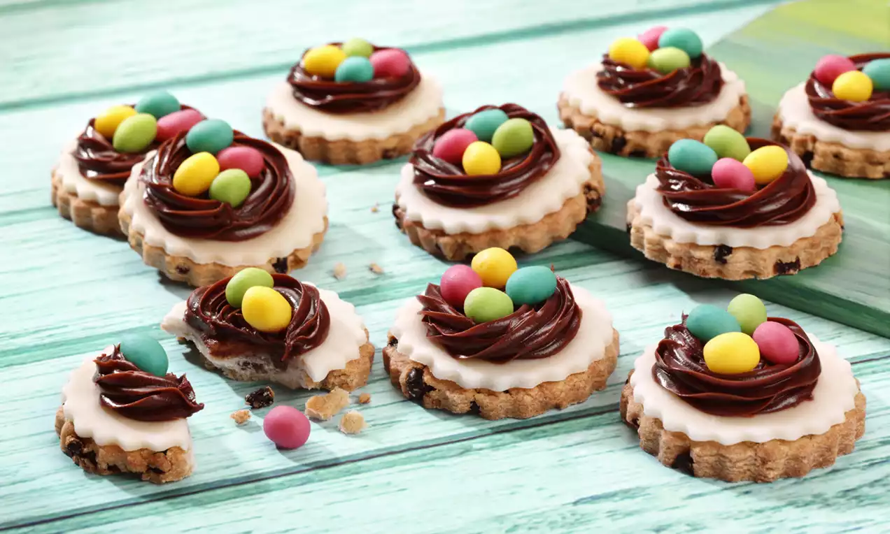 Round biscuits with scalloped edges topped with glossy chocolate nests and colorful mini eggs, arranged on a pale wooden table