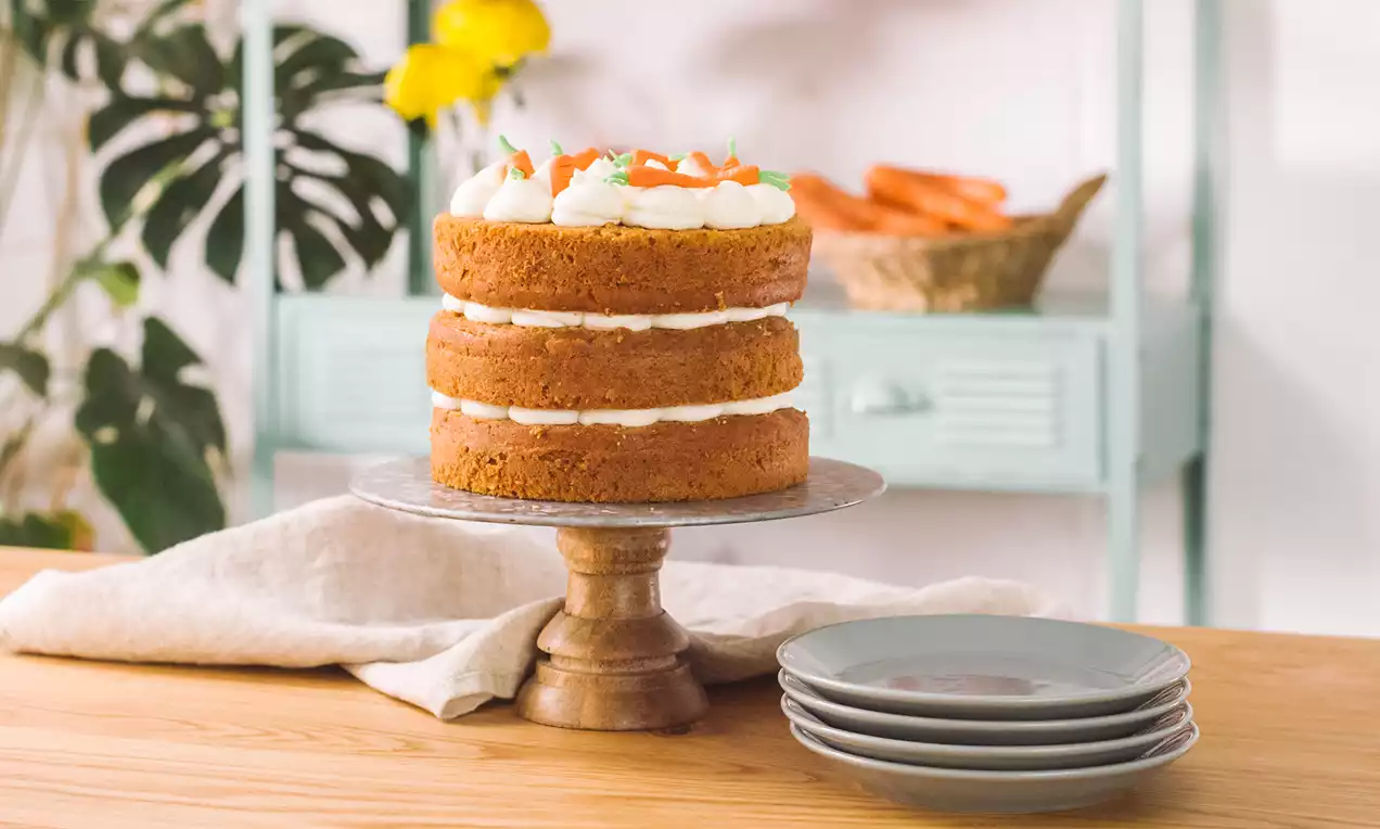 Pastel de capas doradas con relleno cremoso blanco, decorado con crema y zanahoria rallada sobre un pedestal de madera