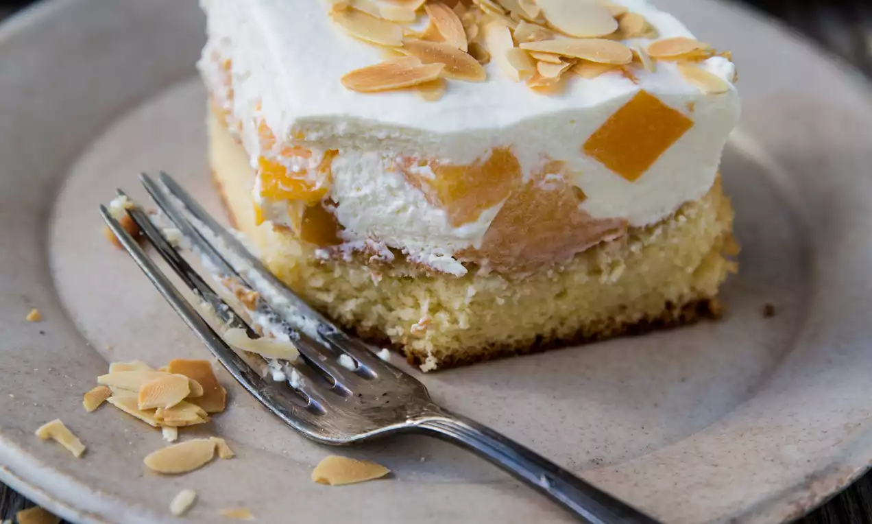Fetta di torta rettangolare su base soffice dorata con strato di crema bianca, pezzi di arancia vivaci e lamelle di mandorle su un piatto rustico
