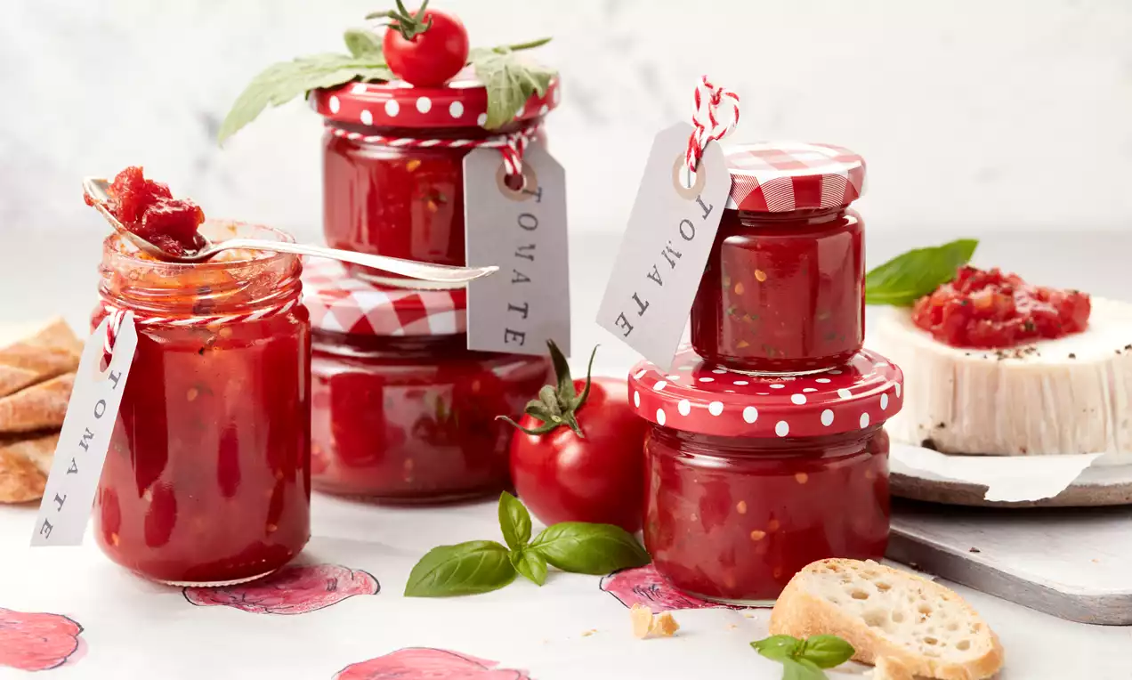 Glänzend rotes Tomaten-Chutney in Gläsern mit gepunkteten Deckeln, garniert mit frischen Tomaten, Basilikum und knusprigem Baguette