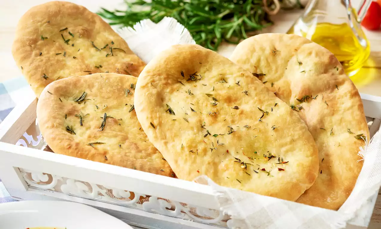 Focaccia dorée et moelleuse parsemée d’herbes, disposée dans un panier blanc doublé de tissu, avec herbes fraîches et huile d’olive à côté