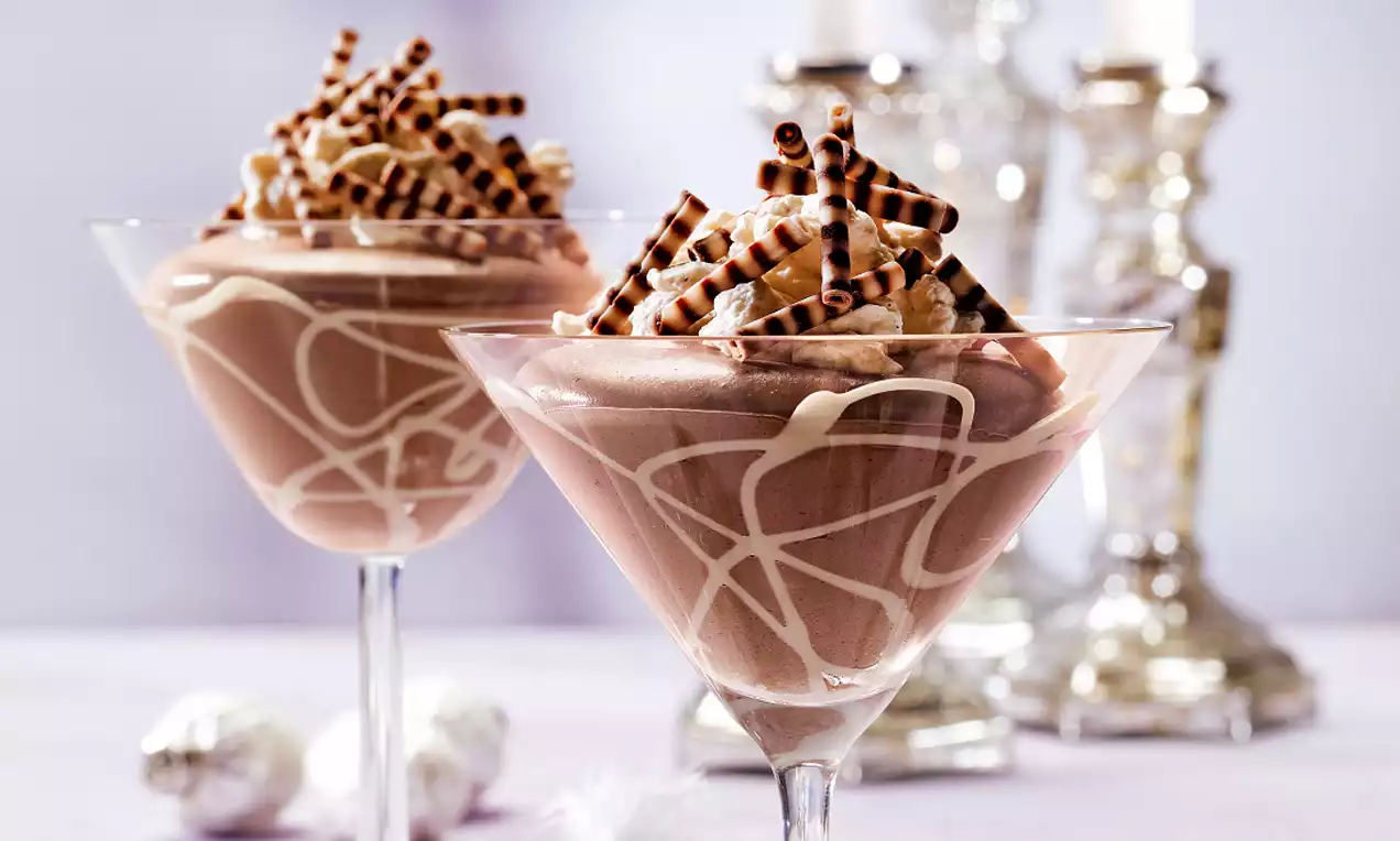 Pharisaeercrème in glazen coupes met linten witte chocolade, bedekt met slagroom en gestreepte chocoladeversiering op een licht paarse ondergrond