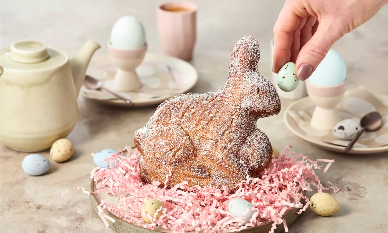 Gebäck-Osterhase aus braunem Teig mit Puderzucker bestäubt, auf rosa Deko-Stroh auf einem Teller, umgeben von bunten Schokoladeneiern