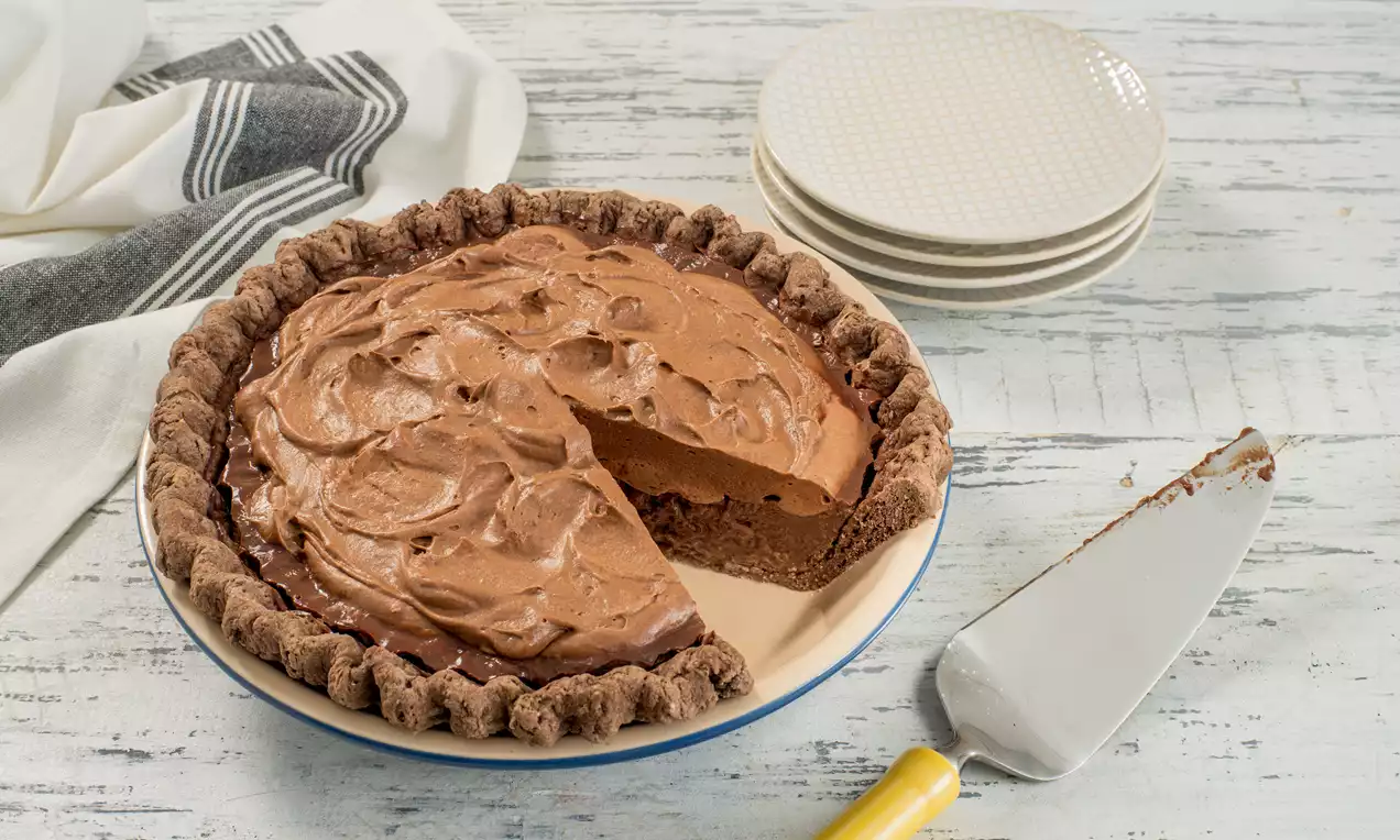 Tarte à croûte brun chocolat garnie d'une mousse chocolatée brillante sculptée en spirales, présentée sur une assiette crème avec une pelle jaunes