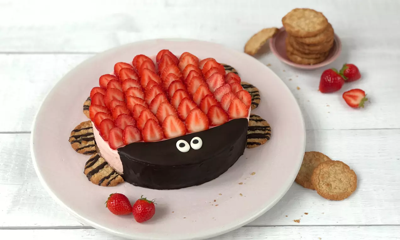 Çilek dilimleriyle kaplanmış, üst kısmı çikolatalı, göz detaylı uğur böceği şeklinde pasta, kurabiye ve çilek süslemeli beyaz tabakta