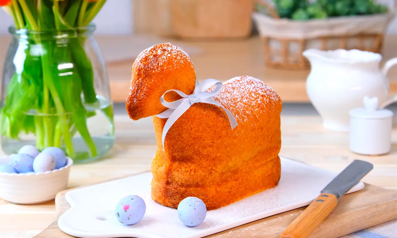 Dolce a forma di agnello dorato con zucchero a velo, rifinito con un fiocco argentato su un piatto bianco, circondato da uova colorate