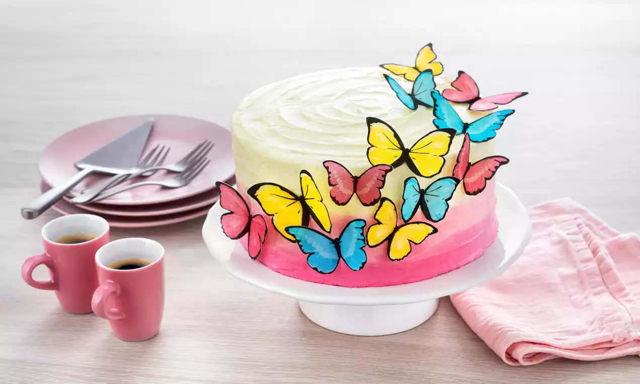 Rund geschichtete Torte mit weiß-rosa Buttercreme und bunten essbaren Schmetterlingen auf weißer Tortenplatte neben rosa Geschirr und Tassen