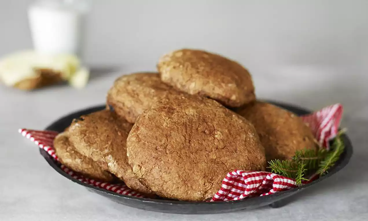 Bruna vorttekakor med rustik textur ligger staplade på en mörk tallrik med rödvitrutigt tyg och grankvist, bakgrund med mjölk och ost