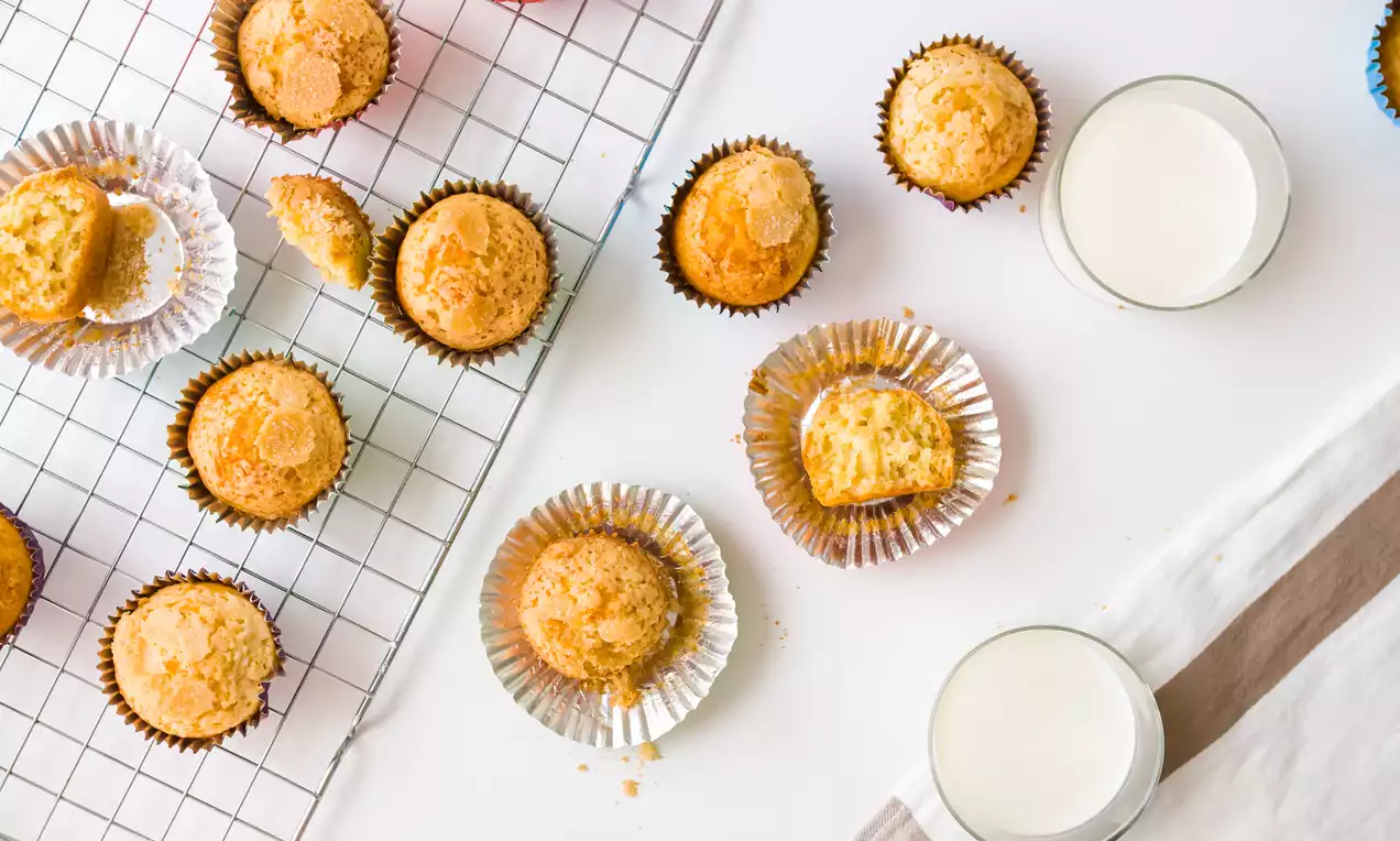 Magdalenas doradas de superficie ligeramente rugosa en cápsulas brillantes sobre rejilla metálica y mesa blanca junto a dos vasos de leche