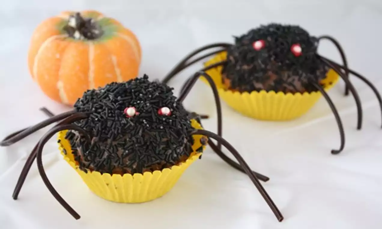 Mustilla nonparelleilla peitetyt hämähäkkimuffinit keltaisissa vuoissa, lakritsi jaloilla ja punaiset silmät, taustalla oranssi kurpitsa