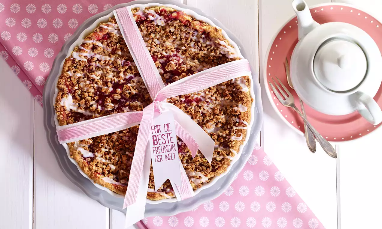 Pflaumenkuchen mit bräunlich-knusprigen Streuseln, in einer runden Form mit rosa Geschenkband dekoriert, auf rosafarbenem Stoff serviert