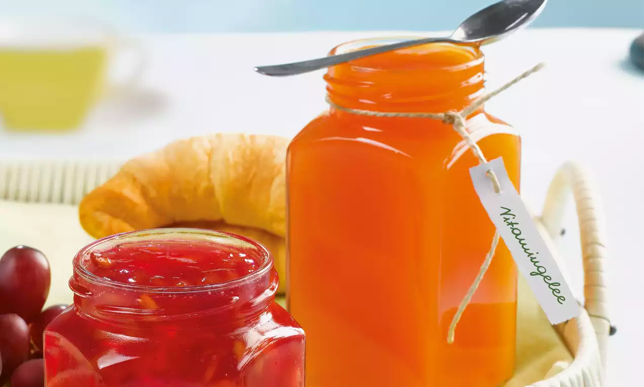 Zwei Gläser mit glänzend rotem Gelee und leuchtend orangefarbenem Gelee, arrangiert mit Weintrauben und Croissant auf heller Platte