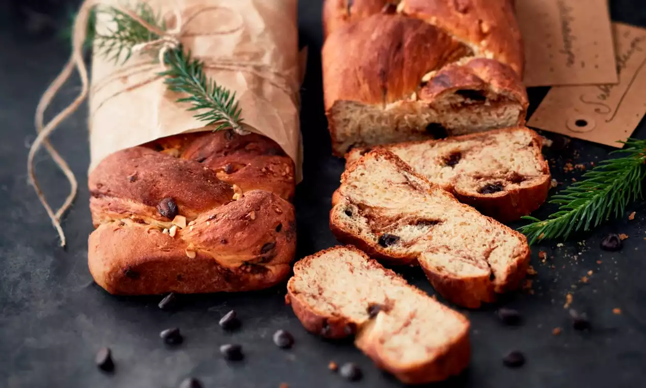 Kullanruskea, kiiltäväpintainen korvapuustinmuotoinen leipä, ilmava sisus ja suklaapaloja, aseteltuna tummajaavaiselle alustalle kuusenhavun kera