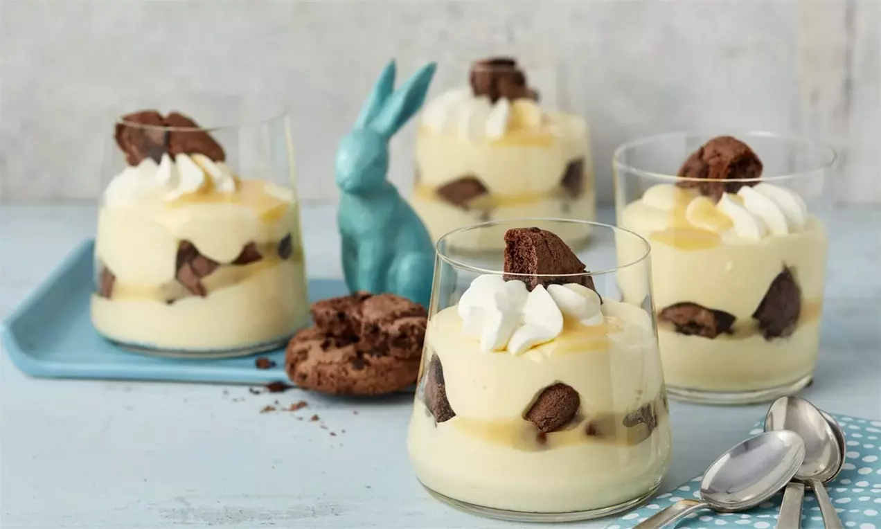 Мек жълт крем в прозрачни чаши с шоколадови парченца, завършен със снежнобяла сметана и сервирано до синя фигурка на зайче