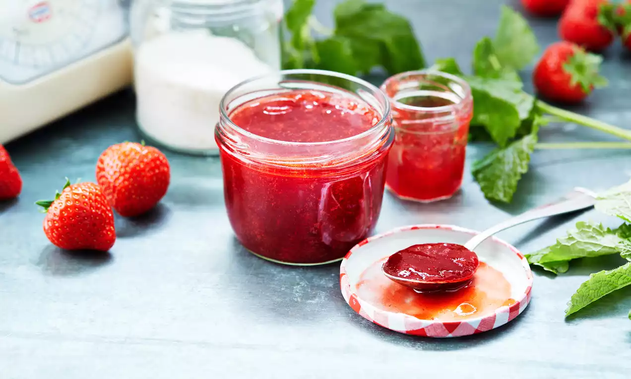 Glanzende aarbeienconfituur in een glazen pot met rode deksel, verse aardbeien en groene blaadjes op een lichtblauwe achtergrond