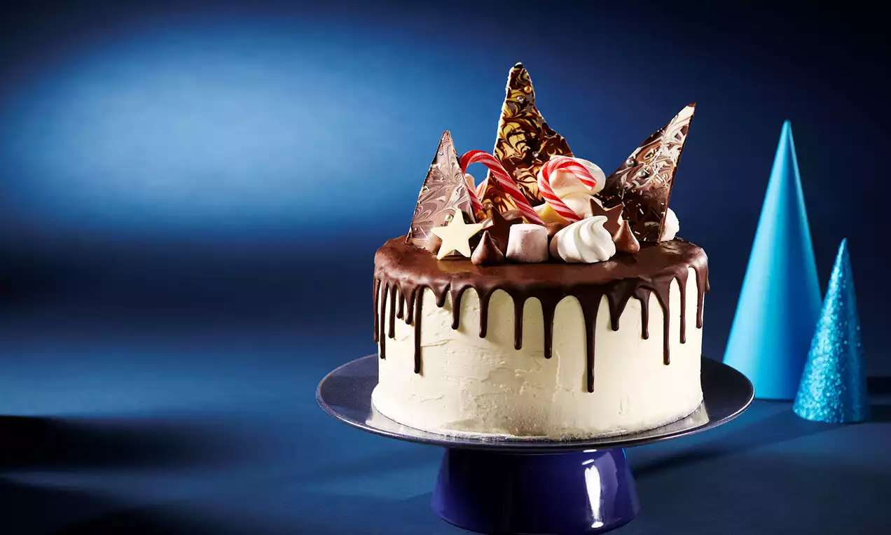 Chocolate-covered cake with smooth white icing, dark chocolate ganache drips, decorated with candy canes, meringues, and chocolate shards on a blue stand