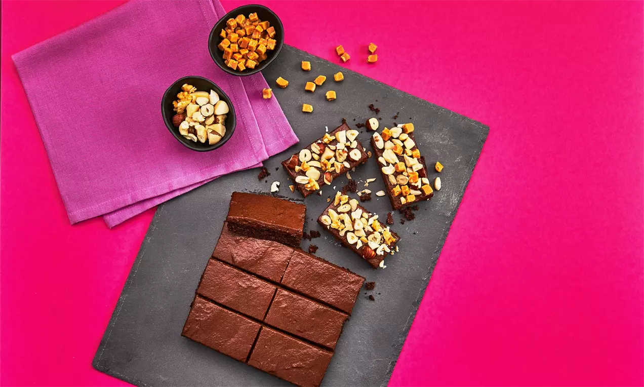 Temno rjavi kvadratni brownie kosi z gladko površino, posuti z mešanico rumenih koščkov in oreščkov na črni ploščici