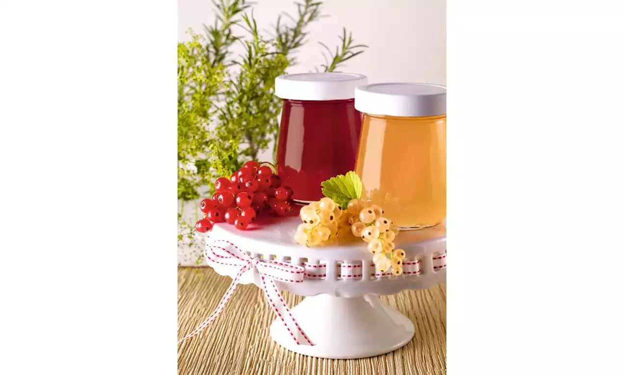 Sklenice rybízového želé s leskle červenou a zlatavou barvou na bílém talíři, zdobené kuličkami rybízu a větvičkami rozmarýnu