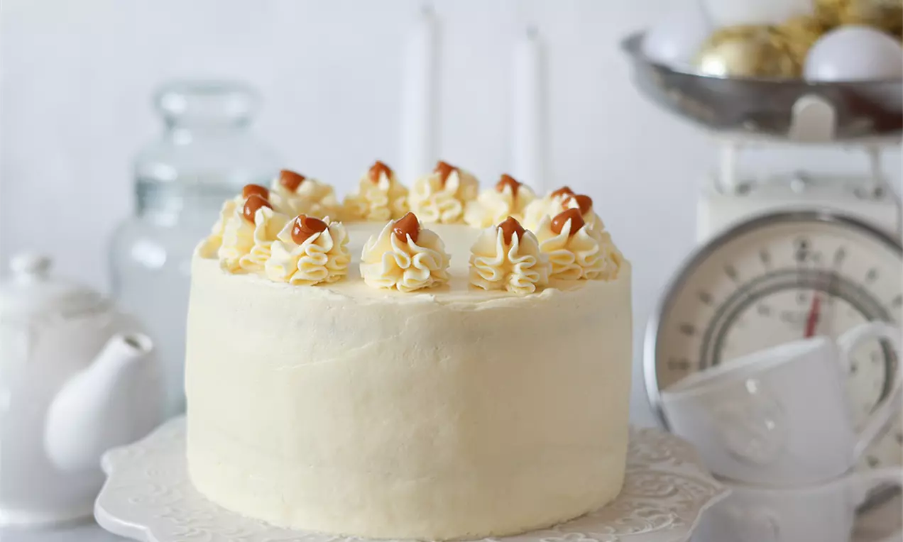 Светла орехова торта с гладко покритие от крем сирене, украсена с рошави върхове крем, завършени с капки солен карамел върху изящна стойка