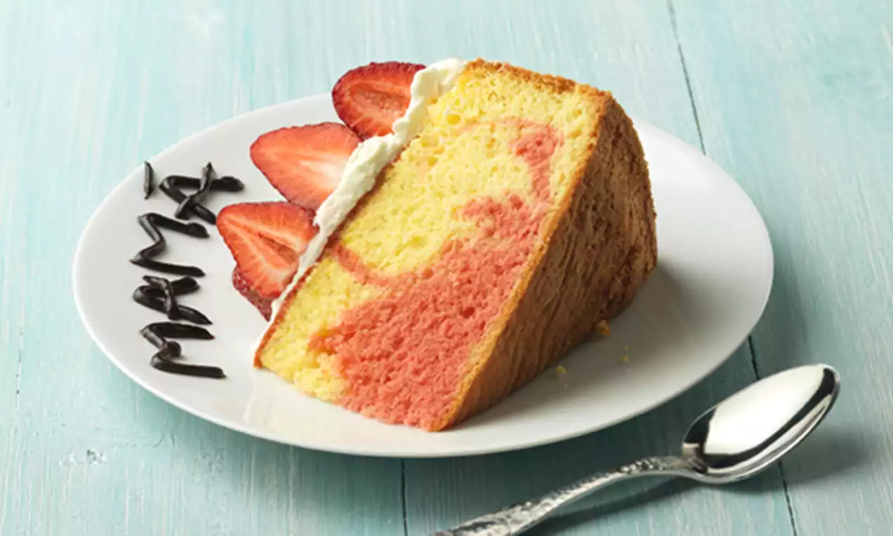 Porción de tarta dorada con textura tierna, en plato blanco decorado con zigzags de chocolate oscuro y una cuchara plateada sobre fondo azul