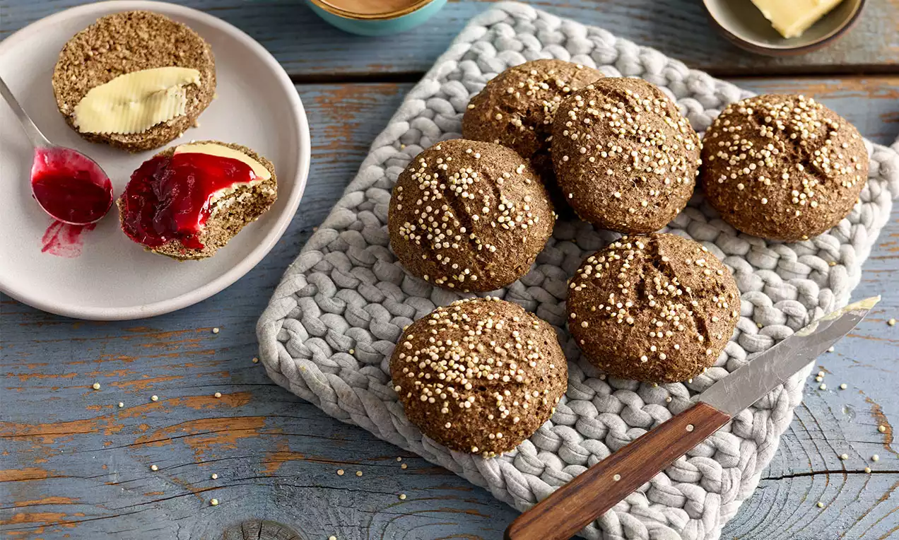 Runde, goldbraune Low-Carb-Brötchen mit luftiger Textur und heller Körnchenstreu auf grauem Untersetzer, daneben Messer und belegte Hälften