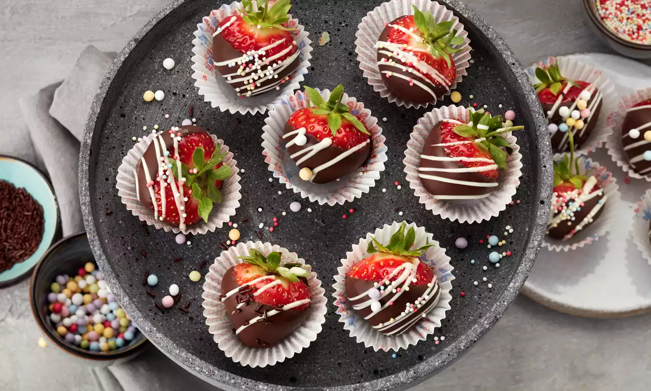 Frische Erdbeeren mit glänzender Schokoladenglasur und weißem Zuckerguss verziert, einzeln in helle Papierförmchen auf schwarzem Teller angerichtet