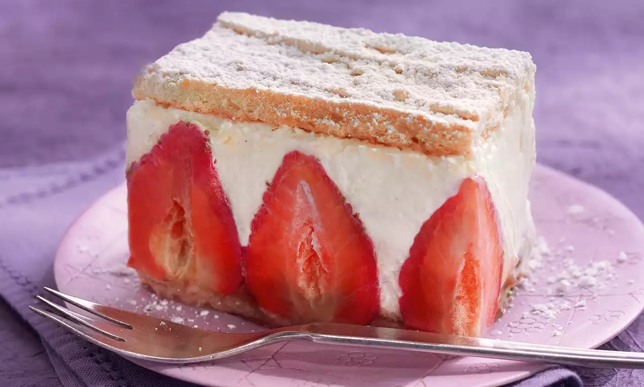 Rechteckiges Dessertstück mit hellen Mürbteig-Schichten, cremiger Füllung und halbierten roten Erdbeeren auf rosa Teller, bestäubt mit Puderzucker