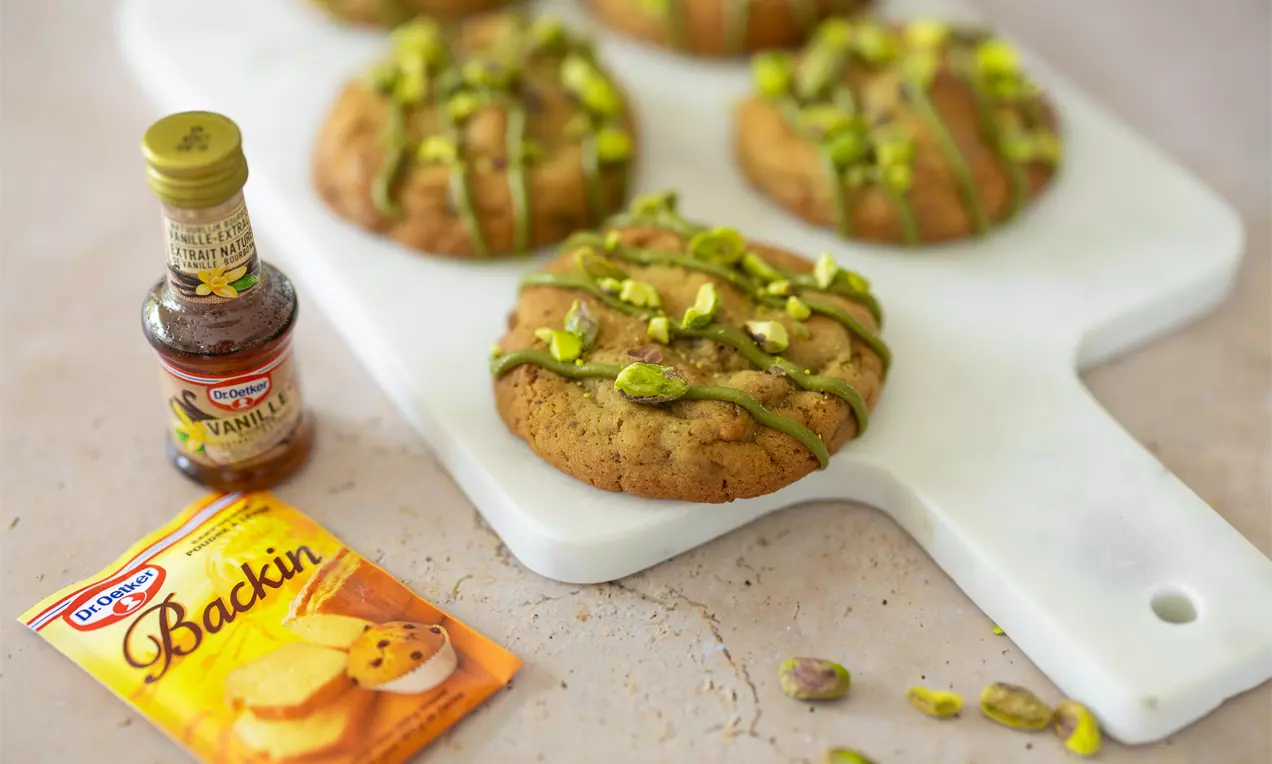Ronde goudbruine koekjes met groene pistachestukjes en glacering, liggend op een wit snijplankje met bakproducten ernaast