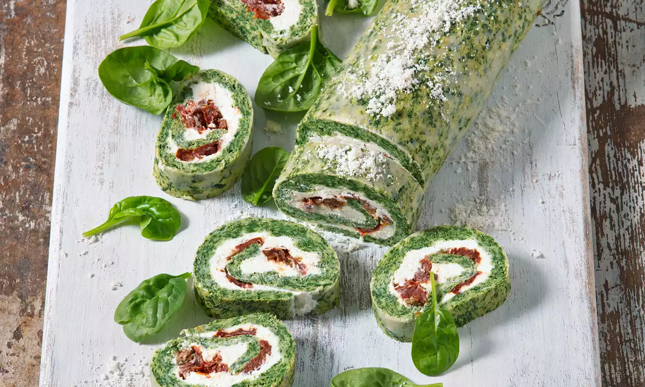 Špenátová roláda s jasně zelenou nadýchanou strukturou, bílou náplní a sušenými rajčaty, vyskládaná na dřevěném prkénku s čerstvými lístky špenátu