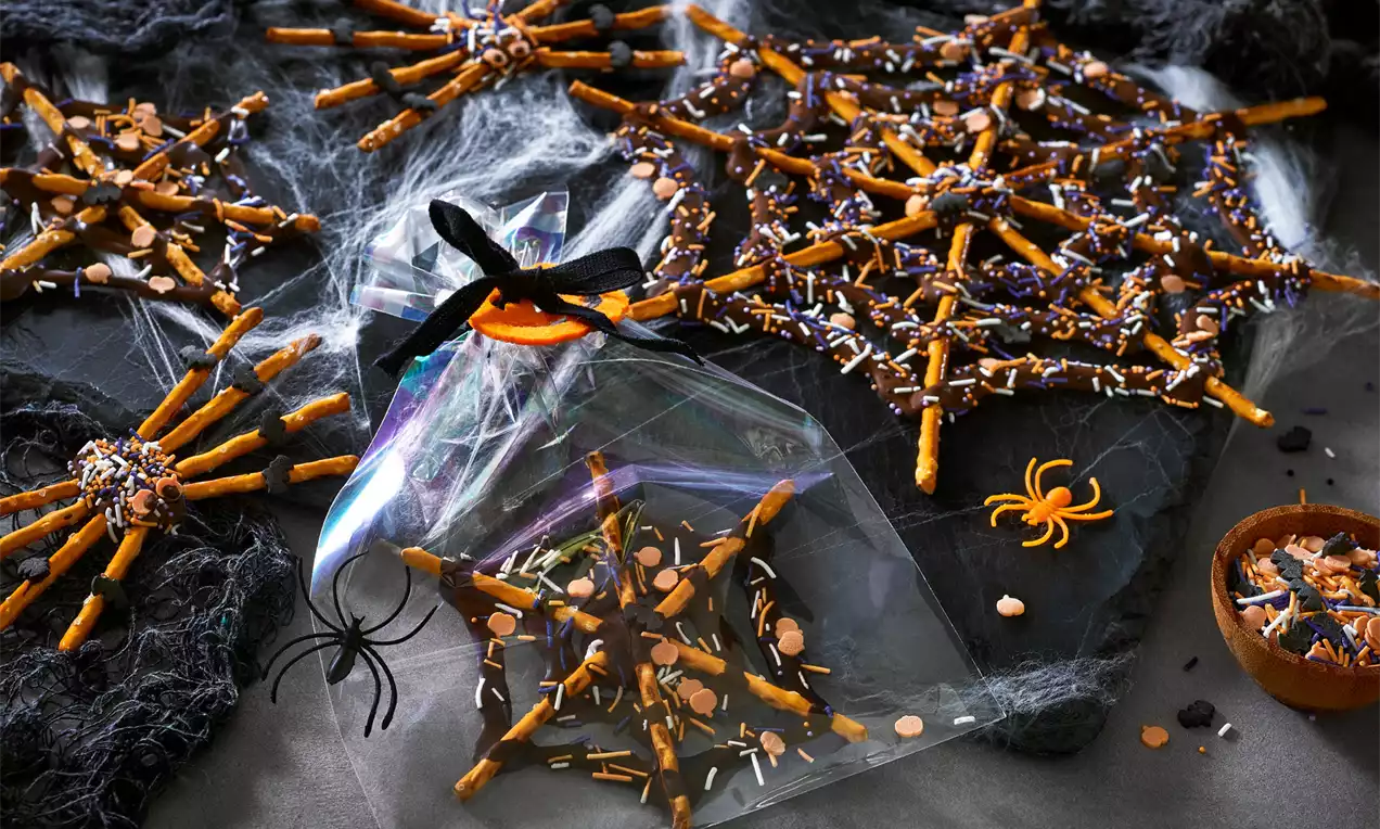 Pânze de păianjen din covrigei aurii lipiți cu ciocolată neagră, presărate cu ornamente colorate, pe fundal de material negru și păianjeni decorativi