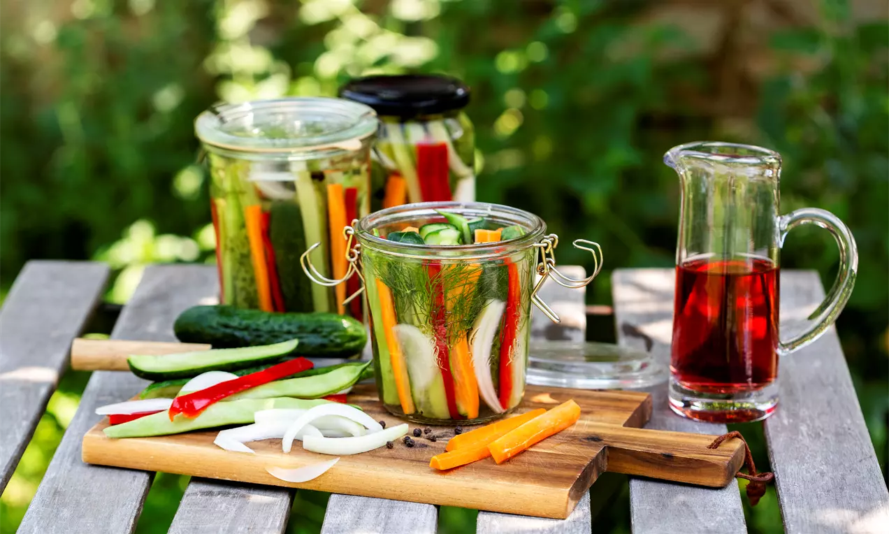 Хрупкави разноцветни зеленчуци и кисели краставички, подредени в буркан с прозрачен капак, заобиколени от дървени плоскости и зеленина