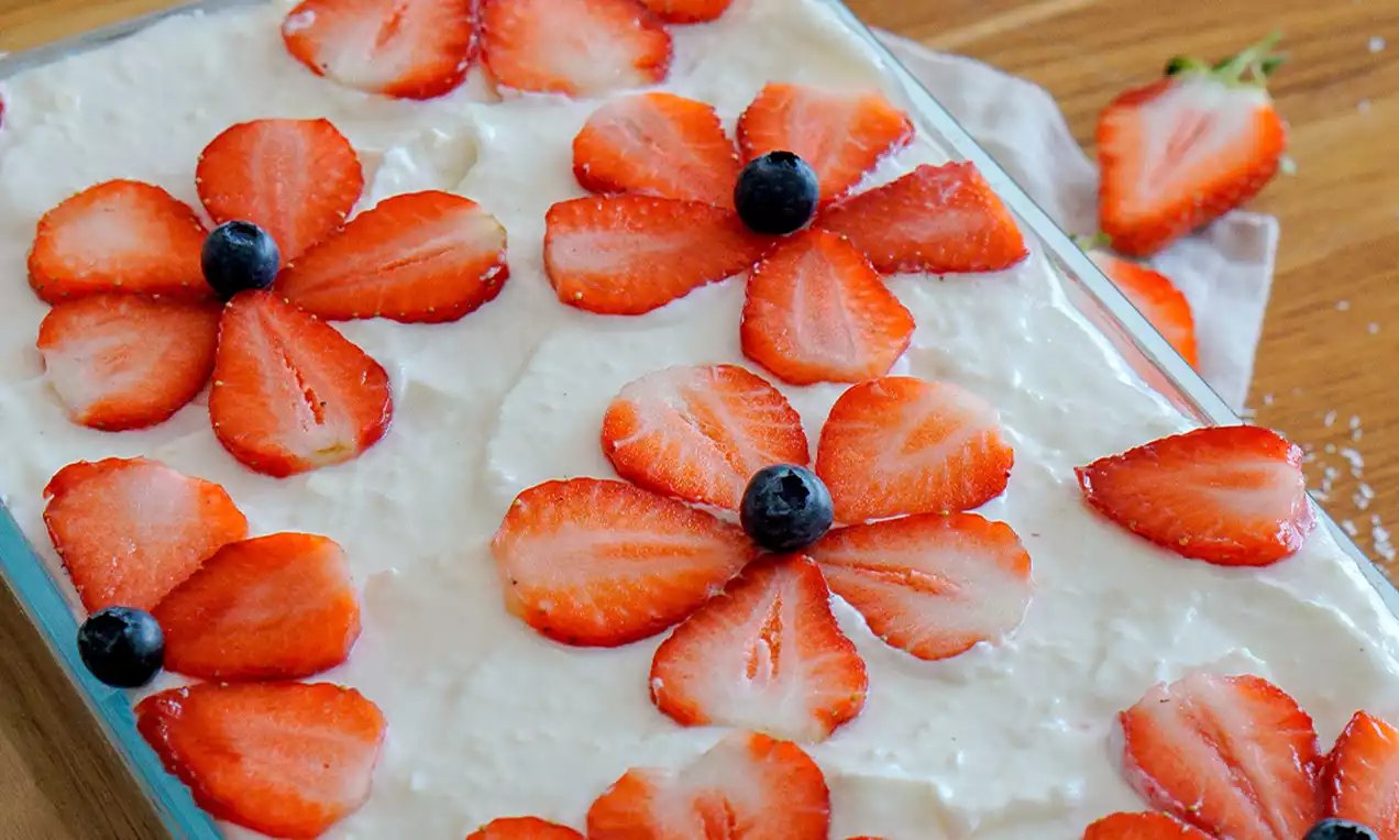 Erdbeer-Kokos-Tiramisu in rechteckiger Glasform, mit weißen Creme-Schichten, dekoriert mit Erdbeerblüten und einzelnen Blaubeeren