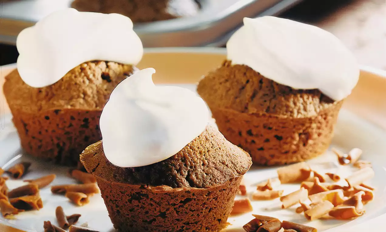 Dunkelbraune Muffins mit lockerer, luftiger Struktur und weißem Sahnehäubchen, dekoriert mit Schokospänen auf einem beigefarbenen Teller