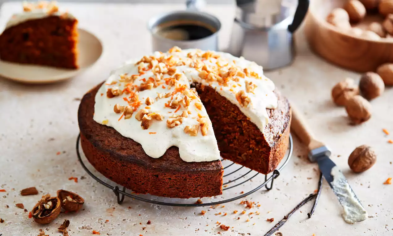 Carrot cake moelleux, nappé de glaçage vanillé blanc, décoré de noix concassées et éclats de carottes sur une grille avec une part servie