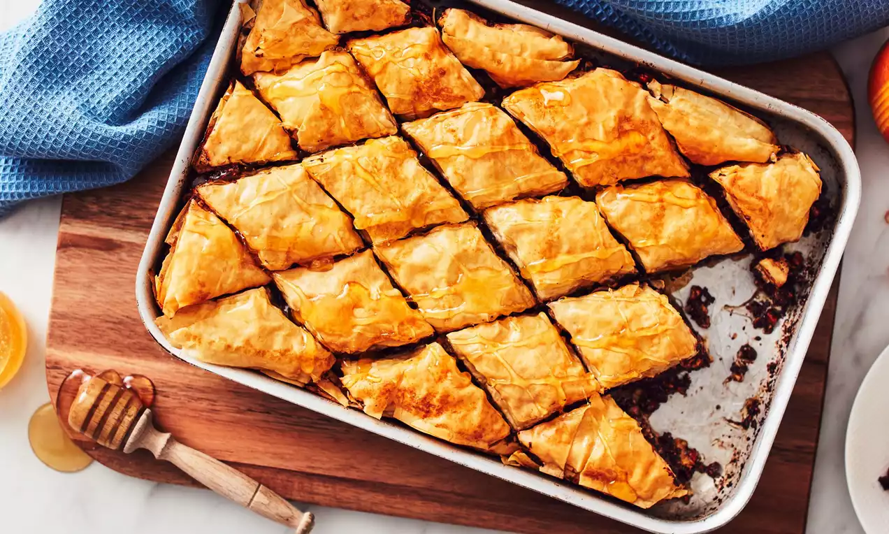 Golden diamond-shaped baklava with flaky layers and dark filling arranged neatly in a white baking tray on a wooden board