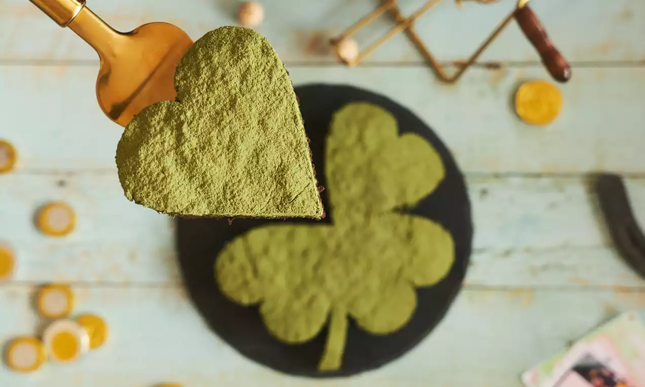 Tarta en forma de trébol recubierta de polvo verde mate con textura fina sobre base negra y una porción en forma de corazón elevada con espátula dorada