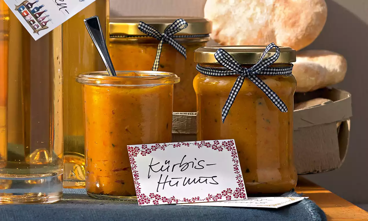 Humus s bundevom u staklenkama zlatno-narančaste boje s kremastom teksturom, ukrašen poklopcima s trakom na plavom stolnjaku