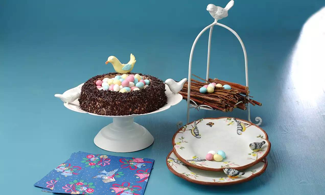 Çikolata kaplı yuvarlak pastanın üstü renkli şeker yumurtalarla süslenmiş, kuş figürüyle sergilenen tabakta çam dalı dekorlu