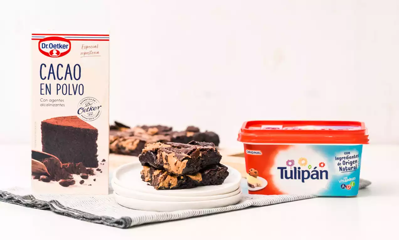 Brownies veganos cuadrados de intenso color marrón, textura densa con vetas de cacahuete en un plato blanco sobre tela gris